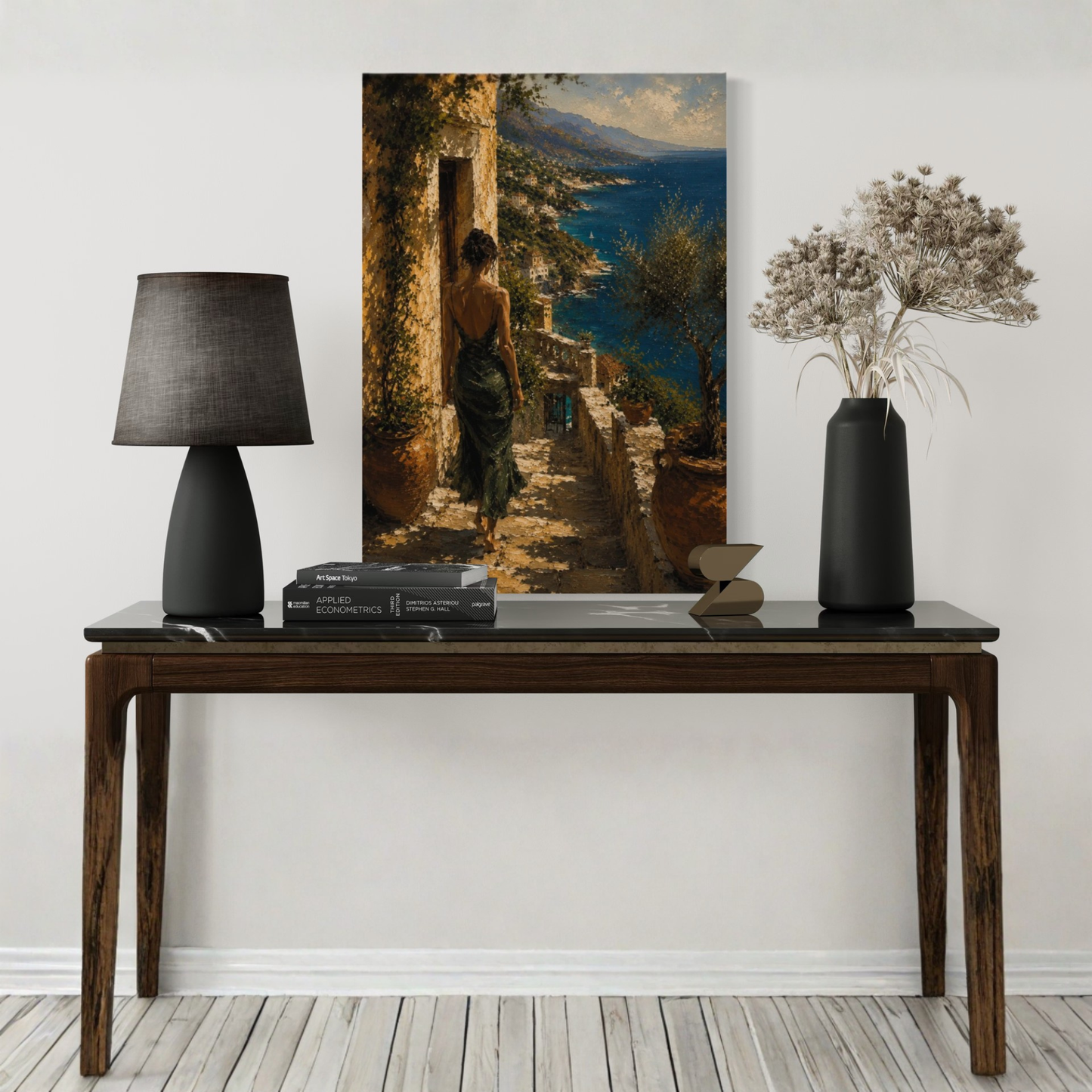 Decorative table with a lamp, books, and a vase in front of a large Mediterranean wall art of a woman by the sea.