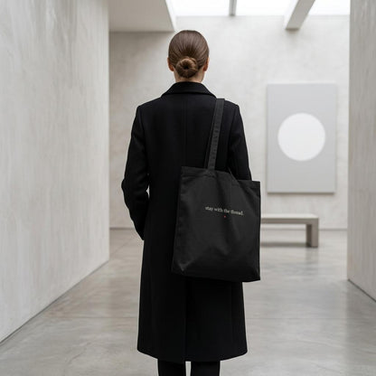 Person in a black coat carrying THE THREAD tote through a minimalist gallery interior.