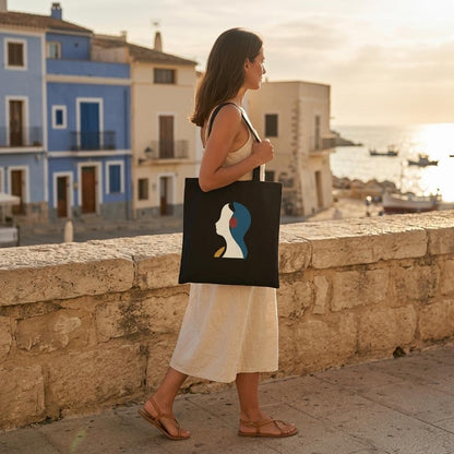 Woman carrying THE LISTENING black cotton tote beside a stone wall in a Mediterranean coastal setting.